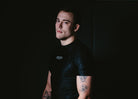 Man wearing a black t-shirt with visible tattoos sitting against a dark background